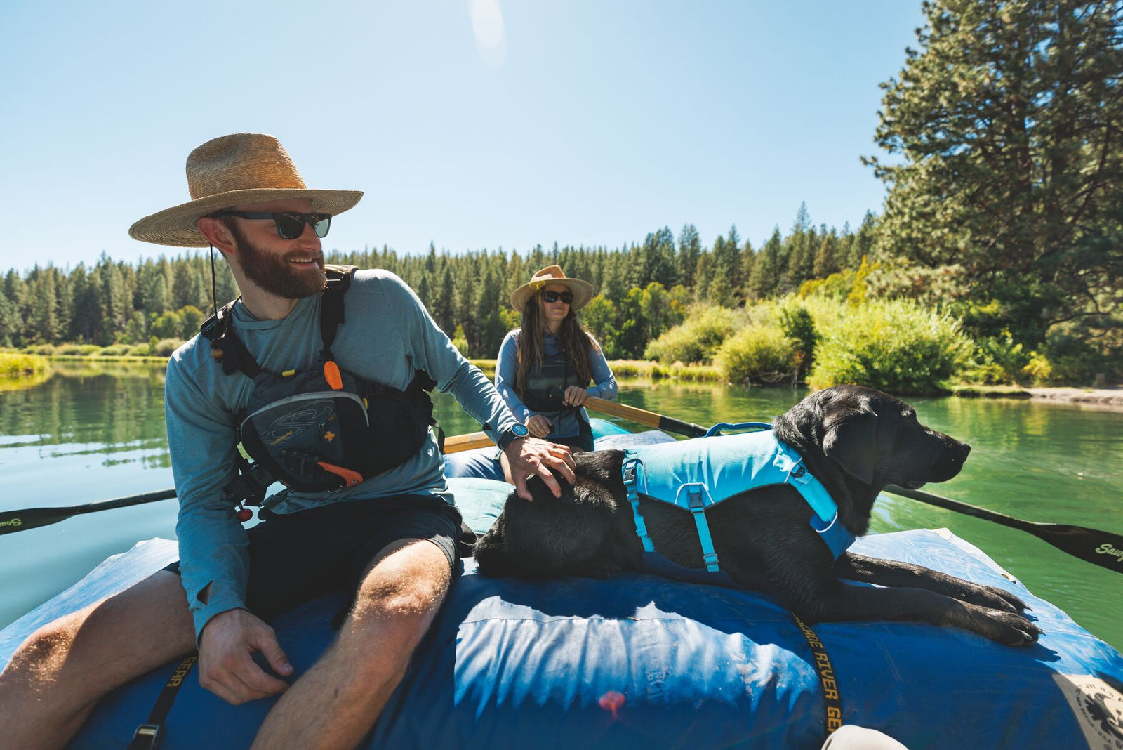 Black dog wearing the Ruffwear Confluence dog life jacket in Biolumin Blue lying on a raft on a calm river, with a man and woman enjoying a day on the water.