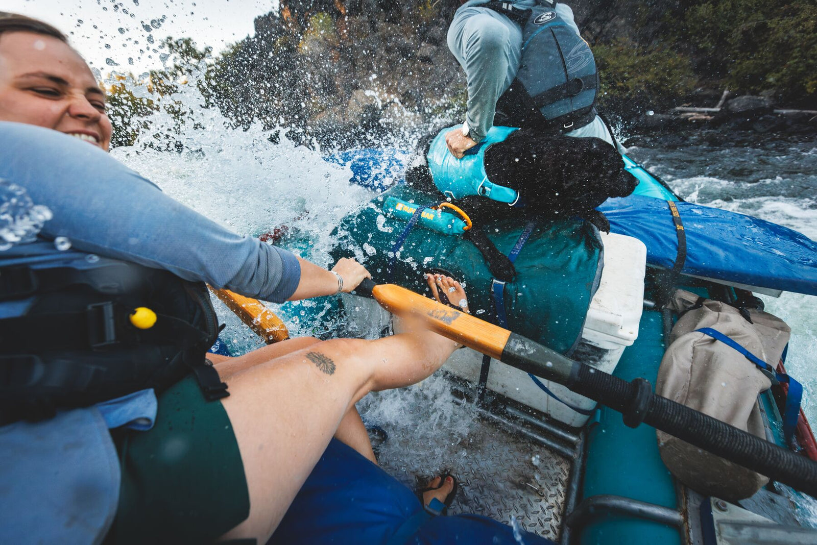 Black dog wearing the Ruffwear Confluence dog life jacket in Biolumin Blue riding a raft through whitewater rapids with two paddlers.