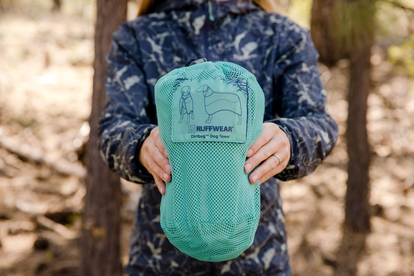 Handler holding the Ruffwear Dirtbag™ Dog Towel packed into its teal mesh stow bag in a pine woodland setting, Ruffwear Dirtbag™ Dog Towel label visible on the front of the bag