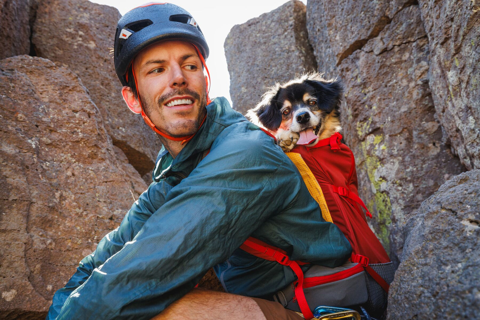Smiling climber wedged in a narrow rock chimney looking back over his shoulder with a black and white dog riding in the Ruffwear Hitch Hiker dog backpack carrier in Red Clay, both looking directly at the camera, towering rock walls either side