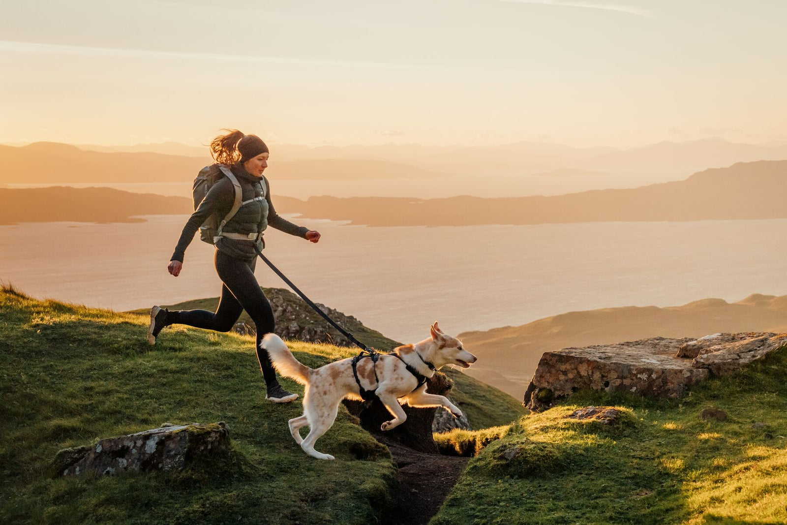 Woman trail running on a hilltop at sunset with a dog on the Ruffwear Ridgeline Lead, loch and mountains in the background