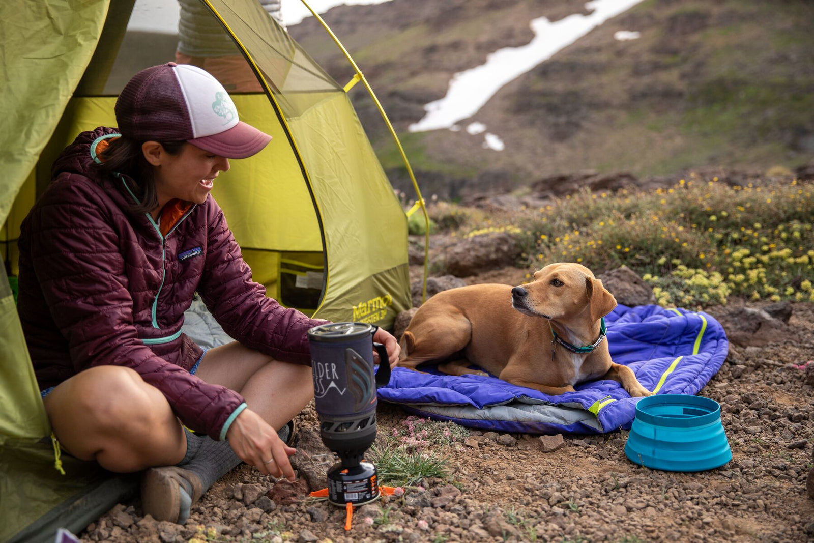 Dog resting on the Ruffwear Highlands Dog Sleeping Bag outside a tent at Steens Mountain, with owner cooking nearby