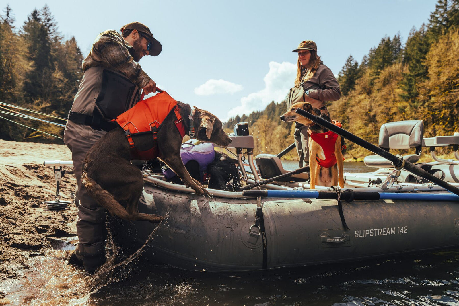 Man lifting a dog wearing the Ruffwear Confluence dog life jacket in Tangelo Orange into a drift boat on a river, with a second dog and woman in the background.