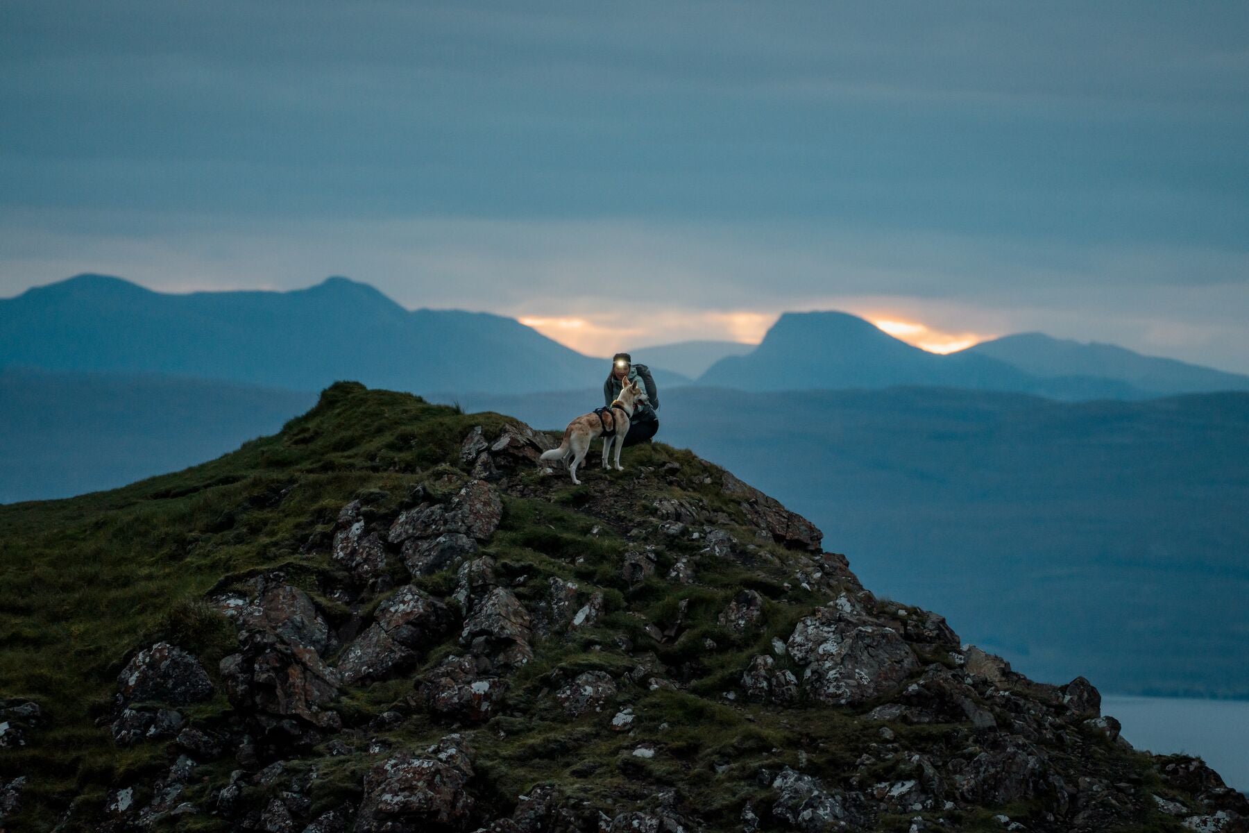 Person and dog sitting together on a mountain summit at dusk wearing the Ruffwear Ridgeline harness, headtorch lit, mountains and water visible in the fading light - built for the adventures that run longer than planned