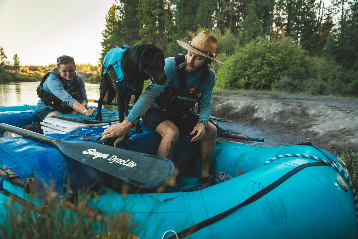 Ruffwear Confluence dog life jacket in Biolumin Blue on a raft at the riverbank, with a woman sat at the back.