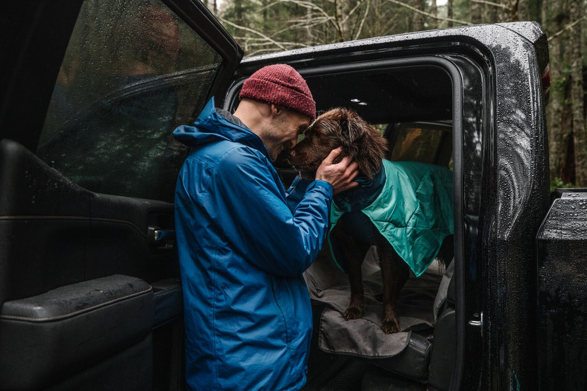 Handler in a blue rain jacket and red beanie leaning in to nuzzle a brown spaniel wearing the Ruffwear Dirtbag™ Dog Towel in Aurora Teal, dog sitting in the open boot of a rain-soaked black 4x4 in a woodland setting — the perfect post-walk moment this towel was made for
