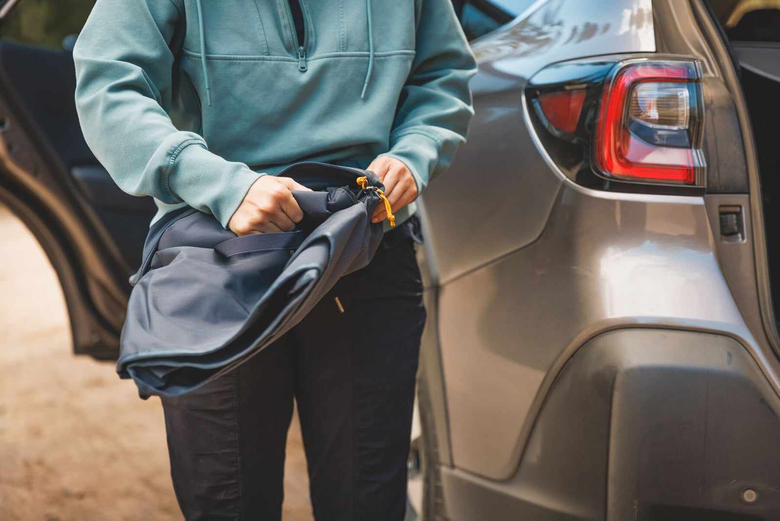 Person in a sage green hoodie unzipping the Ruffwear Dirtbag Seat Cover from its Basalt Gray stow bag beside the rear of a silver SUV in a woodland car park, yellow YKK zip pull visible