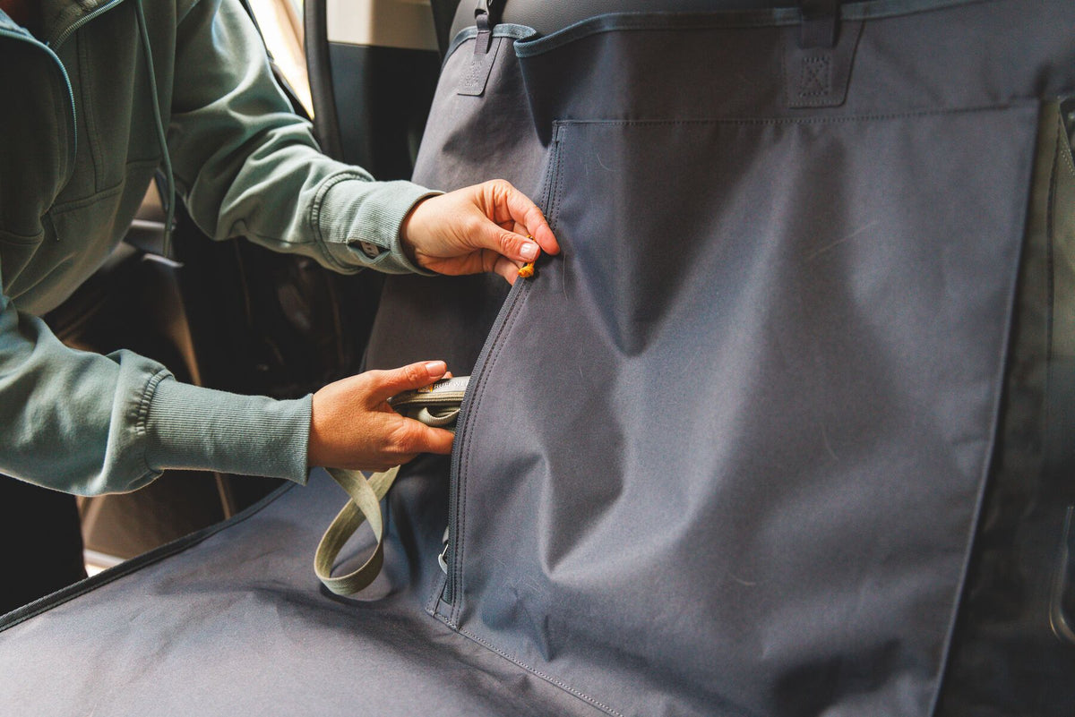 Close-up of hands opening the two-way YKK zip seatbelt access point on the Ruffwear Dirtbag Seat Cover in Basalt Gray, cover fitted across the back seat of a car, stuffer cleat strap visible below