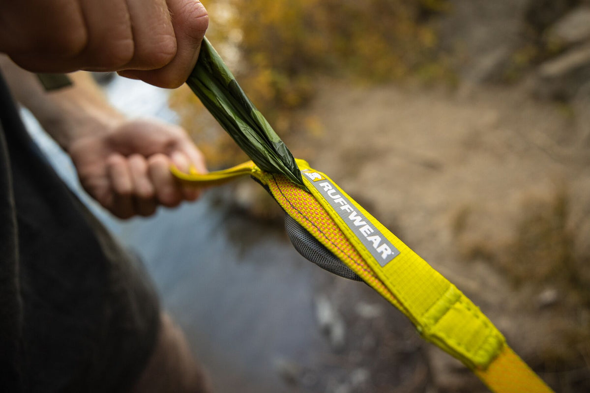 Close-up of a hand pulling a pick-up bag from the stash pocket on the Ruffwear Hi &amp; Light Dog Lead in Lichen Green, outdoors by a stream