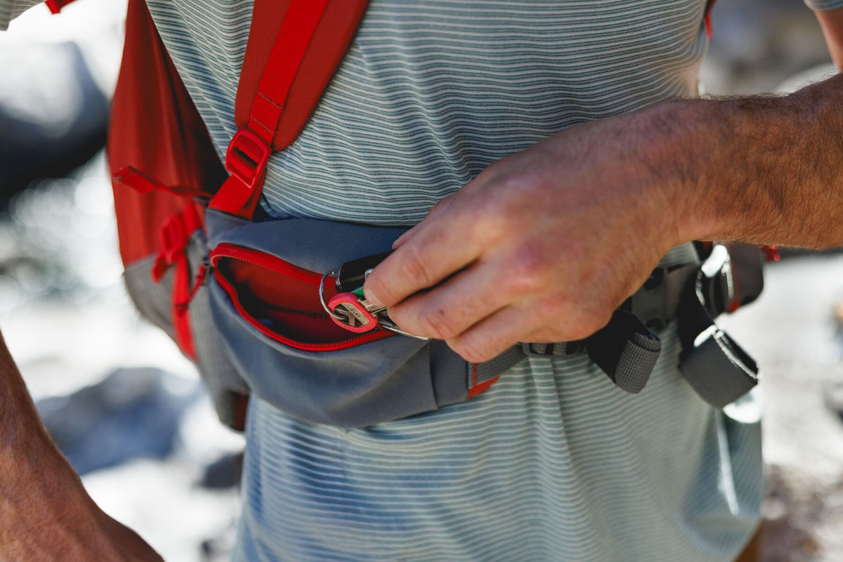 Close-up of hands fastening the hip belt buckle on the Ruffwear Hitch Hiker dog backpack carrier in Red Clay, grey waist strap and red cinch hardware visible against a blurred outdoor background