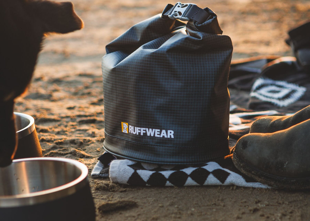 Ruffwear Kibble Kaddie dog food carrier in Basalt Gray sitting upright on a black and white blanket on a sandy beach at golden hour, Ruffwear logo prominent, with a Basecamp Bowl and a dog's nose visible at the edge of the frame