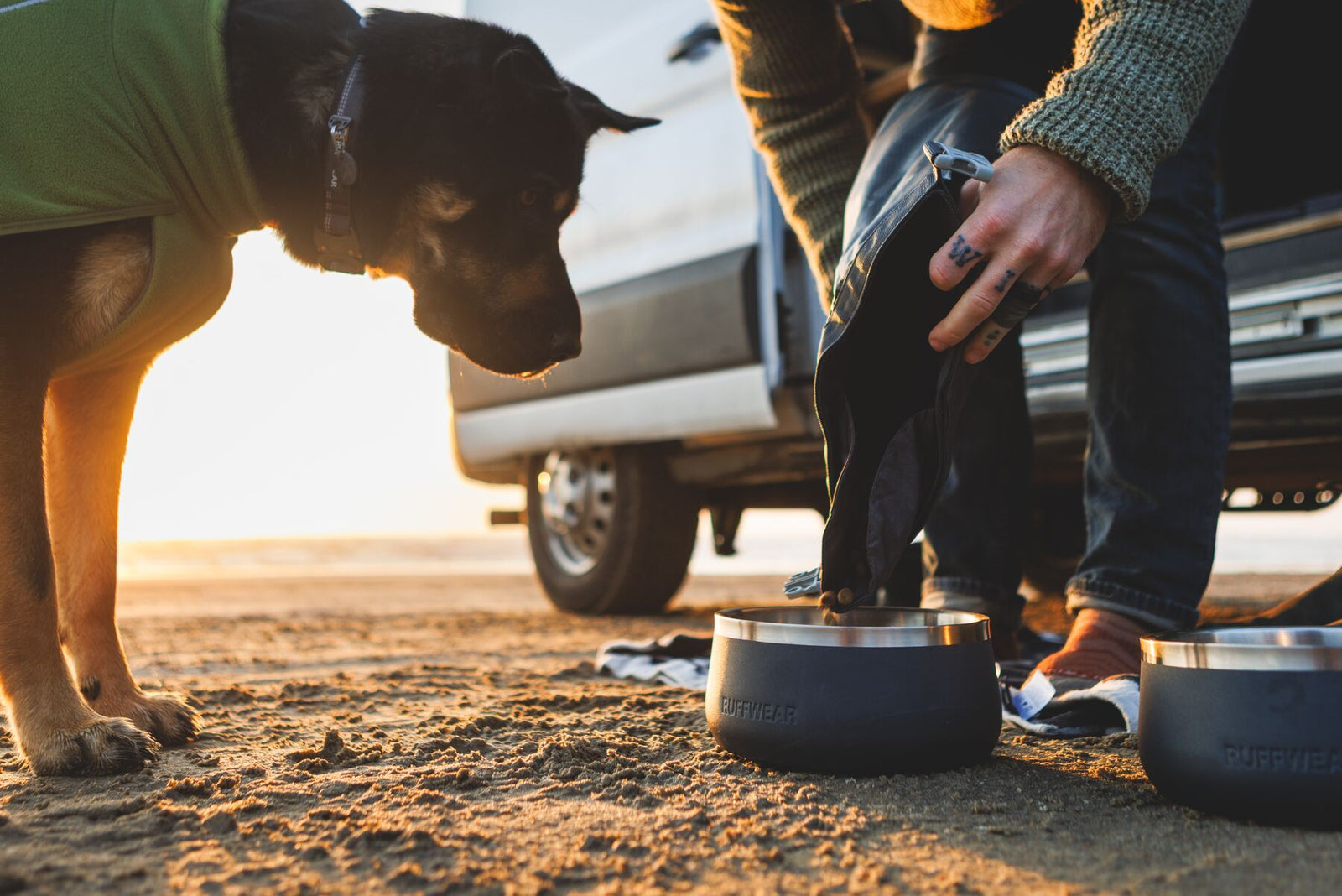 Black dog in a green Ruffwear coat leaning in eagerly as kibble is poured from the Ruffwear Kibble Kaddie into a Ruffwear Basecamp Bowl on a sandy beach at golden hour, campervan and a second bowl visible behind them