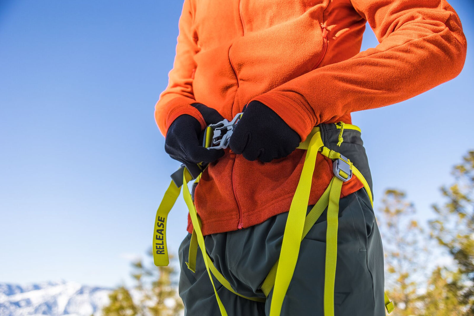 Skier in an orange fleece jacket fastening the Ruffwear Omnijore hip belt in Lichen Green at the waist, gloved hands clicking the central buckle, yellow RELEASE quick-disconnect strap and leg loops visible, snow-covered mountains and pine trees in the background