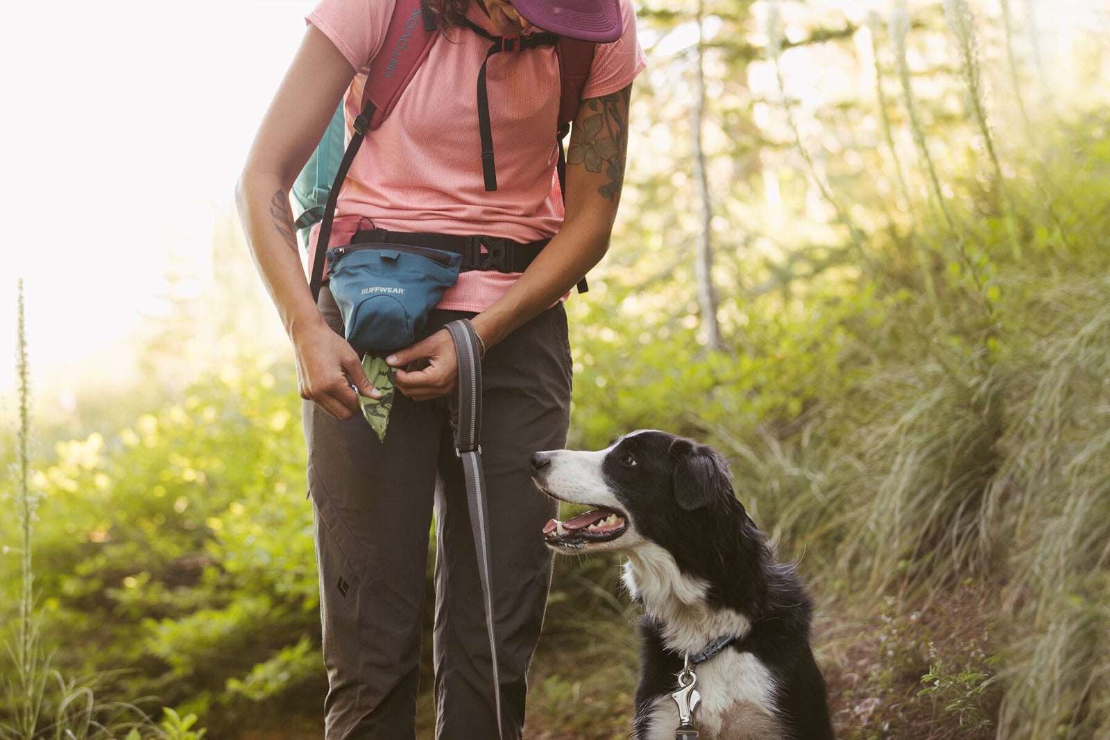 Woman hiker on a sunlit forest trail stowing a dog waste pick-up bag into the Ruffwear Pack Out Bag worn on a waist belt, a black and white border collie sitting at her feet looking up at her, pink backpack and dappled green woodland behind them