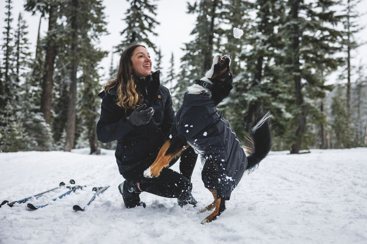A woman kneeling in deep snow throws a snowball while her dog leaps up to catch it, wearing the Ruffwear Powder Hound Coverall in Obsidian Black - snow-covered pine forest in the background, ski poles visible to the left.