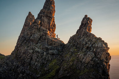 Person and dog picking their way along a narrow rocky ridge at sunset, towering basalt rock formations rising around them, warm golden light catching the stone - the Ruffwear Ridgeline harness built for exactly this