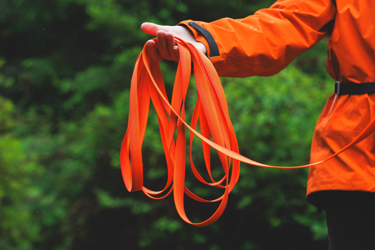 Person in an orange jacket holding a loose bundle of the Ruffwear Rogue Long Line in Blaze Orange against a dark green forest background, showing the flat handleless webbing gathered in one hand