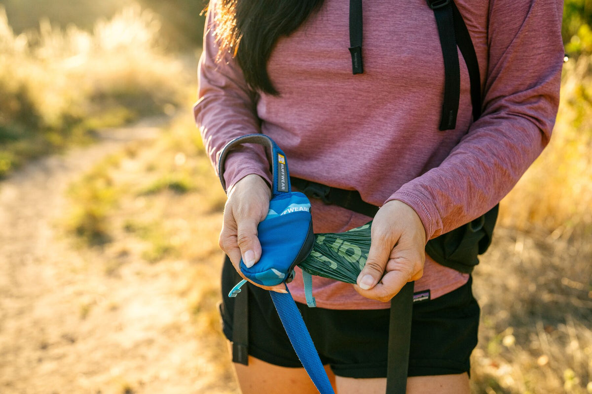 Woman on a sunlit trail pulling a green dog waste pick-up bag from the Ruffwear Stash Bag Mini in Coastal Mountains attached to a blue leash at waist height, backpack straps visible, warm golden grassland behind her
