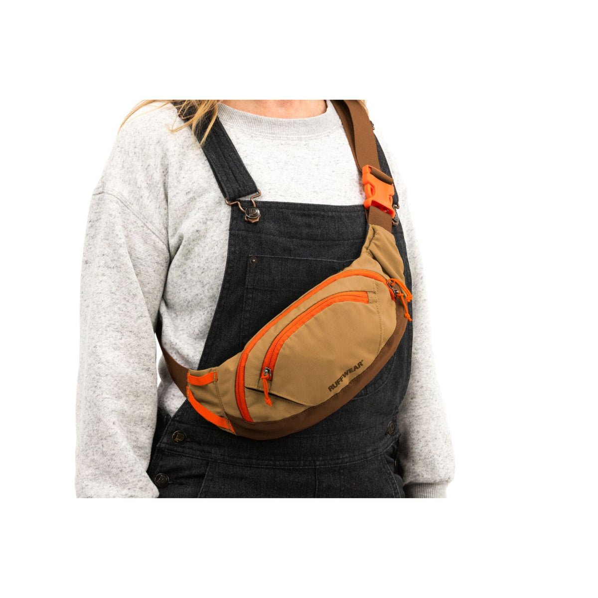Woman wearing the Ruffwear Home Trail hip pack in Brown Bear as a crossbody bag over a white jumper and denim dungarees on a white background, orange zip pulls and side-release buckle visible