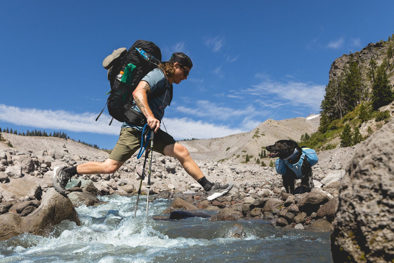 Hiker crossing a mountain stream using trekking poles with black Labrador wearing Ruffwear Palisades Pack in Glacier Lake waiting on rocks, Mount Hood landscape in background