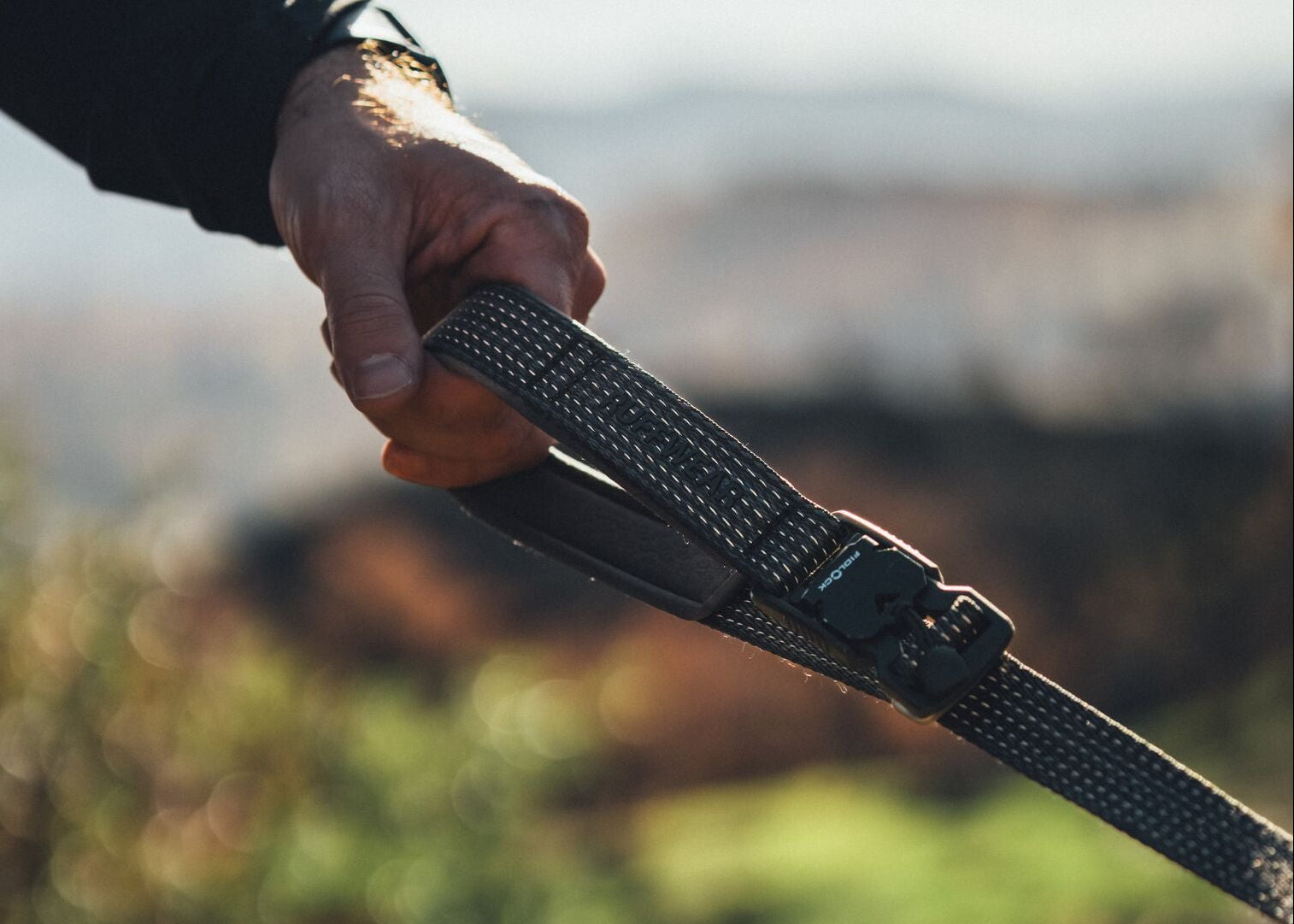 close-up of hand gripping the padded handle of the Ridgeline Lead outdoors, reflective webbing visible, dog blurred in background: