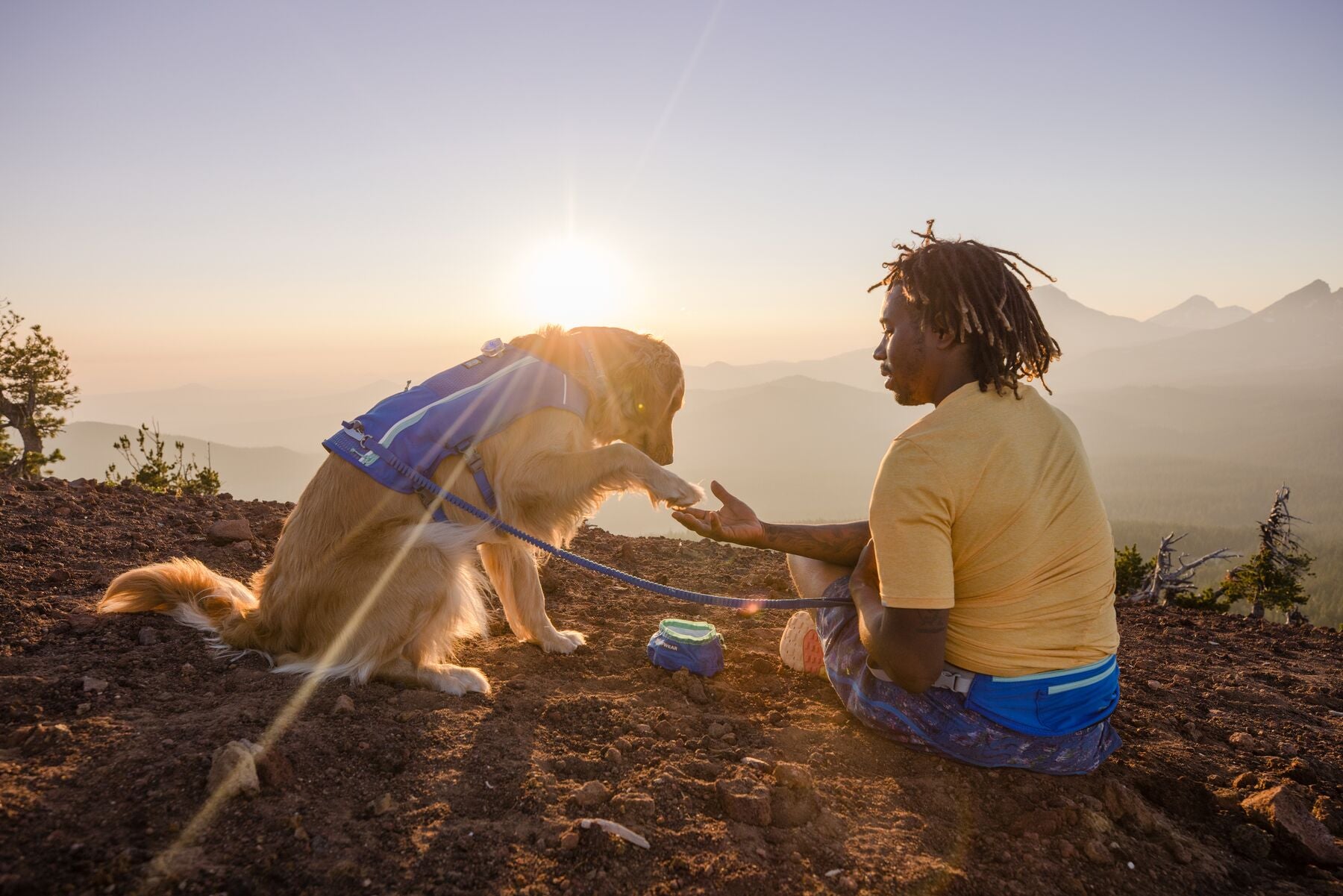 Person sitting with a golden retriever on a mountain summit at sunset, Ruffwear Trail Runner Bowl on the ground beside them